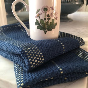 May include: A white ceramic mug featuring a botanical illustration of daisies with green leaves and pink-tipped petals, resting on a folded dark blue textured kitchen towel with a woven grid pattern in a light yellow-green color.