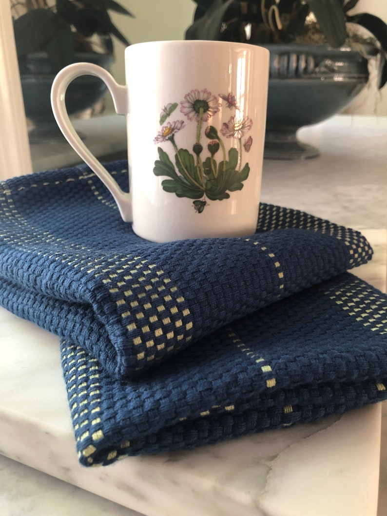 May include: A white ceramic mug featuring a botanical illustration of daisies with green leaves and pink-tipped petals, resting on a folded dark blue textured kitchen towel with a woven grid pattern in a light yellow-green color.