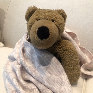 Puede incluir: Un oso de peluche marrón con una nariz negra y una manta rosa claro. La manta tiene un patrón sutil. El oso está sentado en un sofá color crema.