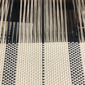 May include: A loom with vertical white and navy blue warp threads extending upward, transitioning into a woven fabric section featuring a repeating checkered pattern of white and navy blue horizontal and vertical threads.