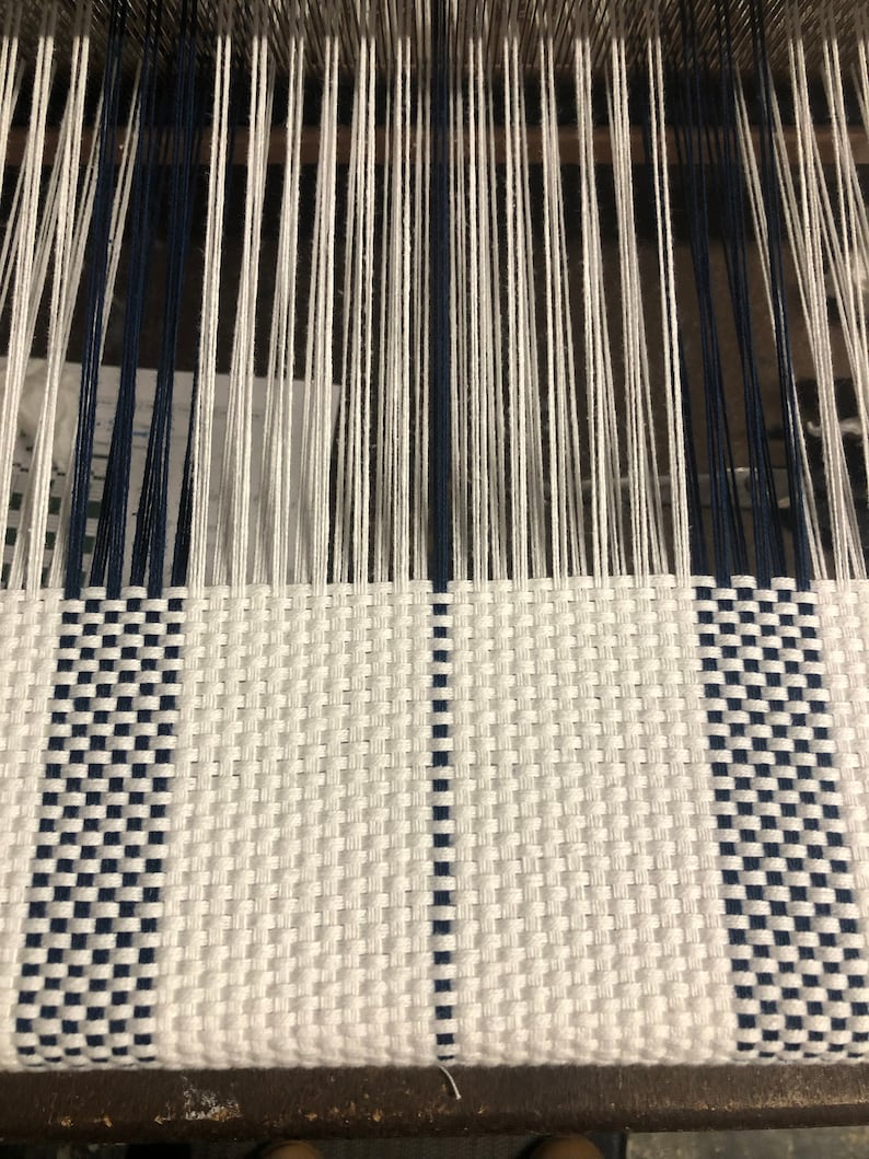 May include: A loom with vertical white and navy blue warp threads extending upward, transitioning into a woven fabric section featuring a repeating checkered pattern of white and navy blue horizontal and vertical threads.
