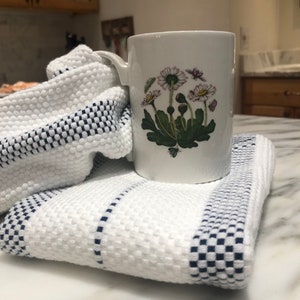 Peut inclure: Une tasse en céramique blanche avec un motif floral de marguerites blanches et de feuilles vertes. La tasse est posée sur une serviette de cuisine rayée blanche et bleue.