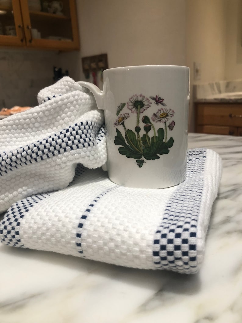 May include: A white ceramic mug featuring a botanical illustration of daisies with green leaves and small insects, resting on a folded white kitchen towel with a textured waffle weave and navy blue checkered stripes along the edges.
