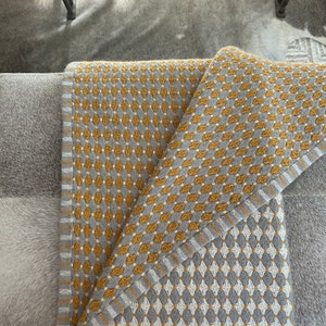May include: A gray and yellow throw blanket with a diamond pattern. The blanket is folded in half and is lying on a gray surface.