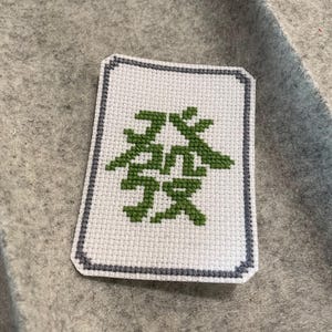 Peut inclure: Une tuile de Mahjong blanche brodée au point de croix avec un caractère vert et une bordure grise. La tuile rectangulaire repose sur une surface grise. Le caractère vert est le mot "Fa" en chinois.