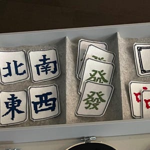 May include: A collection of Mahjong tiles with various Chinese characters in a gray tray. The tiles are white with black, green, and red symbols. The tray is divided into sections, each holding a different set of tiles.