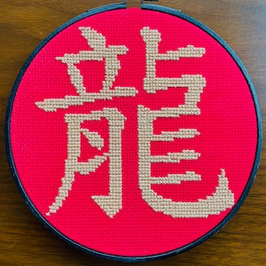 May include: A red circular embroidery hoop with a black frame. The hoop features a white cross-stitch design of the Chinese character for "dragon".