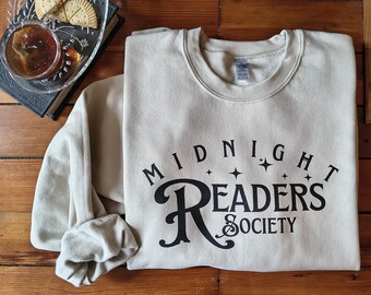 May include: A beige sweatshirt with the text "MIDNIGHT READERS SOCIETY" in black. The sweatshirt is folded on a wooden surface, with a glass dish of crackers and a cup of tea in the background.