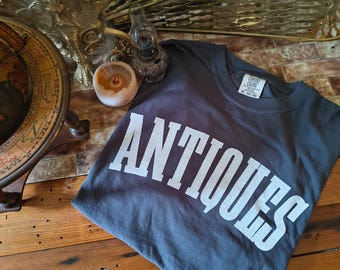 May include: A dark gray t-shirt with the word "ANTIQUES" printed in large white letters. The shirt is folded and lies on a wooden surface. A vintage globe and decorative objects are in the background.