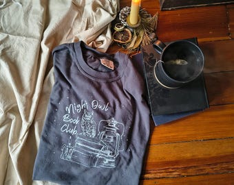 May include: A dark gray t-shirt with the text "Night Owl Book Club" and an owl illustration. The shirt is folded on a wooden surface, next to a lit candle, a black mug, and books. The scene evokes a cozy, literary theme.