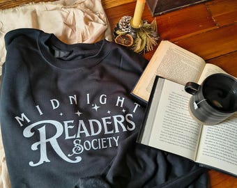 May include: A black sweatshirt with the text "MIDNIGHT READERS SOCIETY" in white script. The sweatshirt is folded next to an open book, a black mug with tea, and a lit candle in a decorative holder. The items are arranged on a wooden surface.
