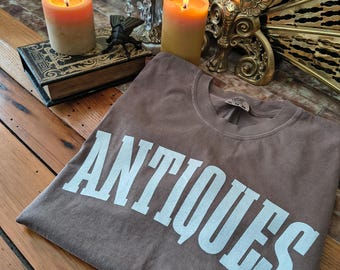 May include: A brown t-shirt with the word "ANTIQUES" printed in large, white, block letters. The shirt is displayed on a wooden surface, with lit candles and a decorative book in the background.