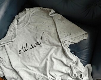 May include: A light gray t-shirt with the words "old soul" in black script. The shirt has a distressed, marbled effect. The t-shirt is folded on a dark blue leather surface.