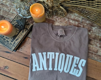 May include: A brown t-shirt with the word "ANTIQUES" in large white block letters. The shirt is folded on a wooden surface, with lit candles and antique decor in the background.