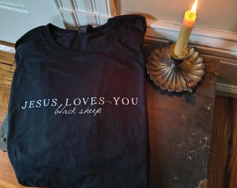 May include: Black t-shirt with the text "JESUS LOVES YOU black sheep" in white script. The shirt is folded on a wooden surface next to a lit candle in a decorative holder.
