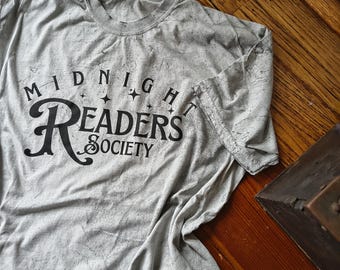 May include: A light gray t-shirt with the text "MIDNIGHT READERS SOCIETY" in black. The shirt has a distressed, vintage look. The text is arched above the word "READERS". The shirt is folded on a wooden surface.