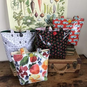 May include: A collection of tote bags with various patterns, including fruits, cherries, mathematical symbols, and retro campers. Each bag features a button closure and a contrasting interior lining. The bags are displayed on a wooden surface with a vintage suitcase and a botanical print in the background.