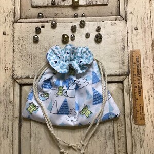 Op de afbeelding: Een witte trekkoordtas met een blauw-wit patroon van een cartoonfiguur. De tas heeft een gerimpelde bovenkant en is gemaakt van stof. De tas ligt op een houten oppervlak.