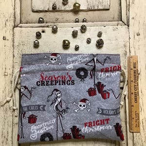 Op de afbeelding: Een grijze trekkoordtas met een kerstthema met Jack Skellington uit The Nightmare Before Christmas. De tas heeft een wit trekkoord en is versierd met rode en zwarte schedels, kransen en de tekst "Season's Creepings", "Frightfully Festive", "Fright Christmas" en "The Chills".