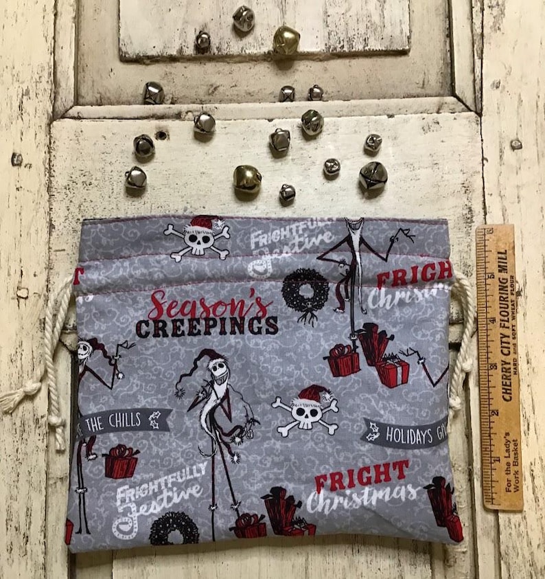Op de afbeelding: Een grijze trekkoordtas met een kerstthema met Jack Skellington uit The Nightmare Before Christmas. De tas heeft een wit trekkoord en is versierd met rode en zwarte schedels, kransen en de tekst "Season's Creepings", "Frightfully Festive", "Fright Christmas" en "The Chills".