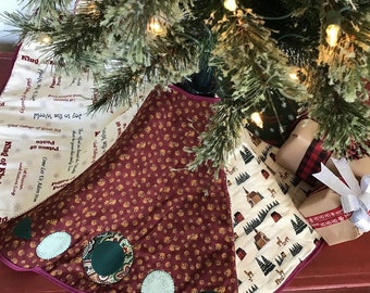 May include: A Christmas tree skirt made of fabric with a patchwork design. The skirt features a burgundy section with a pattern of small gold bells, a cream section with a pattern of small houses and trees, and a cream section with a pattern of snowflakes and the words "King of Kings", "Peace", "Joy to the World", and "Come Let Us Adorn".
