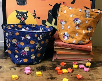 May include: Two fabric trick-or-treat bags with Peanuts characters. One bag is blue with orange pumpkins and the other is orange with white ghosts. Both bags are filled with candy.
