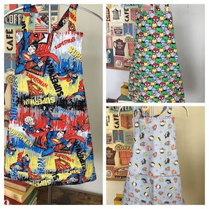 May include: Three different superhero-themed aprons. The first apron is red, yellow, and blue with a Superman pattern. The second apron is green, blue, and red with a Marvel superhero pattern. The third apron is blue with a Batman and Superman pattern.