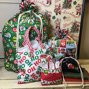 Op de afbeelding: Een set van vier kerstcadeauzakken met een Grinch-thema. De grootste tas is groen met een wit trekkoord en heeft een patroon van de Grinch en kerstversiering. De andere drie tassen zijn kleiner en hebben verschillende Grinch-themapatronen. De tassen zijn allemaal van stof en hebben een trekkoord sluiting.