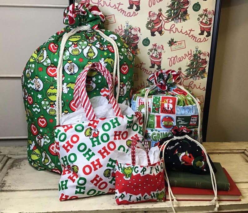 Op de afbeelding: Een set van vier kerstcadeauzakken met een Grinch-thema. De grootste tas is groen met een wit trekkoord en heeft een patroon van de Grinch en kerstversiering. De andere drie tassen zijn kleiner en hebben verschillende Grinch-themapatronen. De tassen zijn allemaal van stof en hebben een trekkoord sluiting.