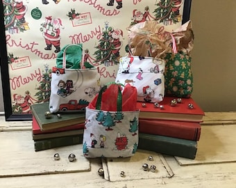 May include: Four small fabric gift bags with handles, featuring a Peanuts theme. The bags are decorated with various Peanuts characters and designs, including Snoopy, Woodstock, and a Christmas tree. The bags are sitting on top of a stack of books with a red, green, and brown color scheme.
