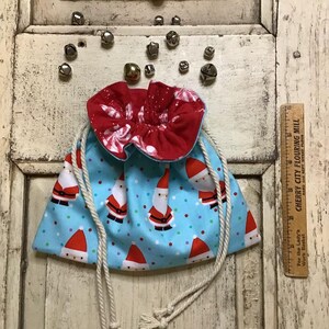 Op de afbeelding: Een blauwe en rode trekkoordtas met een wit stippenpatroon en een print van de kerstman. De tas heeft witte trekkoorden en een gerimpelde rode voering.