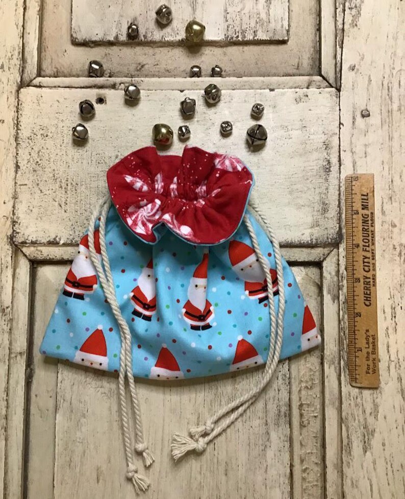 Op de afbeelding: Een blauwe en rode trekkoordtas met een wit stippenpatroon en een print van de kerstman. De tas heeft witte trekkoorden en een gerimpelde rode voering.