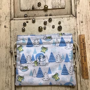 Op de afbeelding: Een witte trekkoordtas met een blauw en groen patroon van een populair fictief personage en kerstboom. De tas is gevuld met kleine zilveren belletjes.