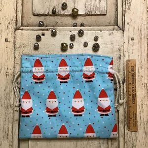 Op de afbeelding: Een blauwe trekkoordtas met een herhalend patroon van kerstmanfiguren en kleurrijke stippen. De tas is gemaakt van stof en heeft een wit trekkoord.