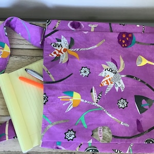 May include: A purple gardening apron with a floral design made of cut-out text and images. The apron has pockets and long ties. A yellow notepad and pens are visible inside the apron.