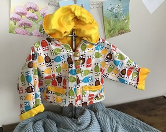 May include: A white hooded raincoat with a yellow hood and a colorful owl print. The raincoat has a yellow trim and yellow buttons.