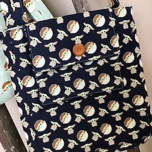 Puede incluir: Una bolsa de mano negra con un patr&oacute;n repetido de un personaje verde con ojos grandes. La bolsa tiene un bot&oacute;n marr&oacute;n y un bolsillo m&aacute;s peque&ntilde;o en la parte delantera.