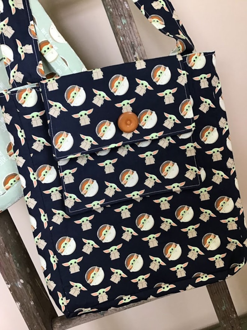 Puede incluir: Una bolsa de mano negra con un patr&oacute;n repetido de un personaje verde con ojos grandes. La bolsa tiene un bot&oacute;n marr&oacute;n y un bolsillo m&aacute;s peque&ntilde;o en la parte delantera.