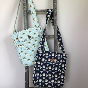 Puede incluir: Dos bolsas de tela con un patr&oacute;n de un personaje de dibujos animados. Una bolsa es azul claro con fondo blanco y la otra es azul oscuro con fondo blanco.