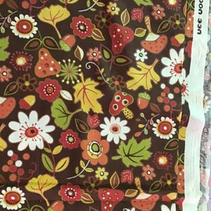 Peut inclure: Tissu marron orné d'un motif floral coloré. Le motif comprend des fleurs blanches, oranges et rouges, des feuilles vertes, des glands et des champignons. Le texte "uee woodland" est visible sur le bord droit.