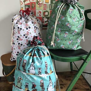 May include: Three drawstring gift bags with holiday-themed patterns. One bag features Mickey Mouse, another has a green pattern with cartoon characters, and the third has a blue pattern with snowman and character designs. Each bag has a white drawstring.