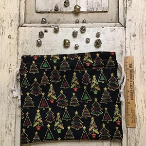 Op de afbeelding: Een zwarte trekkoordtas met een kerstboompatroon. De bomen zijn omrand met goud en hebben rode, groene en gele accenten. De tas is gemaakt van stof en heeft een wit trekkoord.