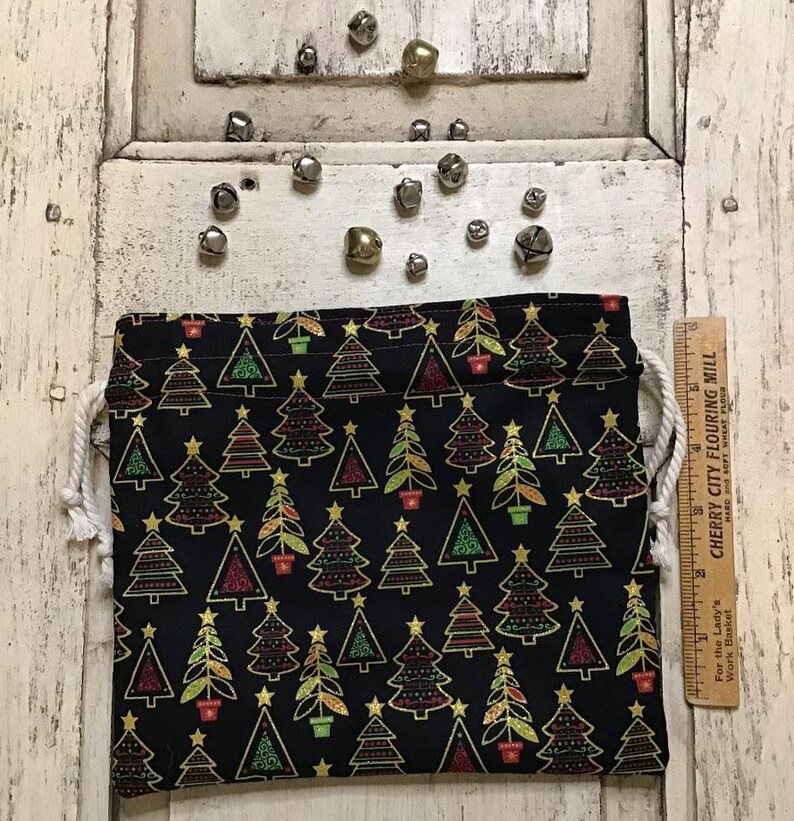 Op de afbeelding: Een zwarte trekkoordtas met een kerstboompatroon. De bomen zijn omrand met goud en hebben rode, groene en gele accenten. De tas is gemaakt van stof en heeft een wit trekkoord.