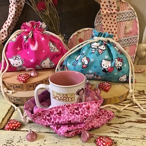 May include: Three pink and blue drawstring bags with Hello Kitty designs, a pink mug with the words "Miss Smarty" on it, and red heart-shaped candies on a wooden surface.