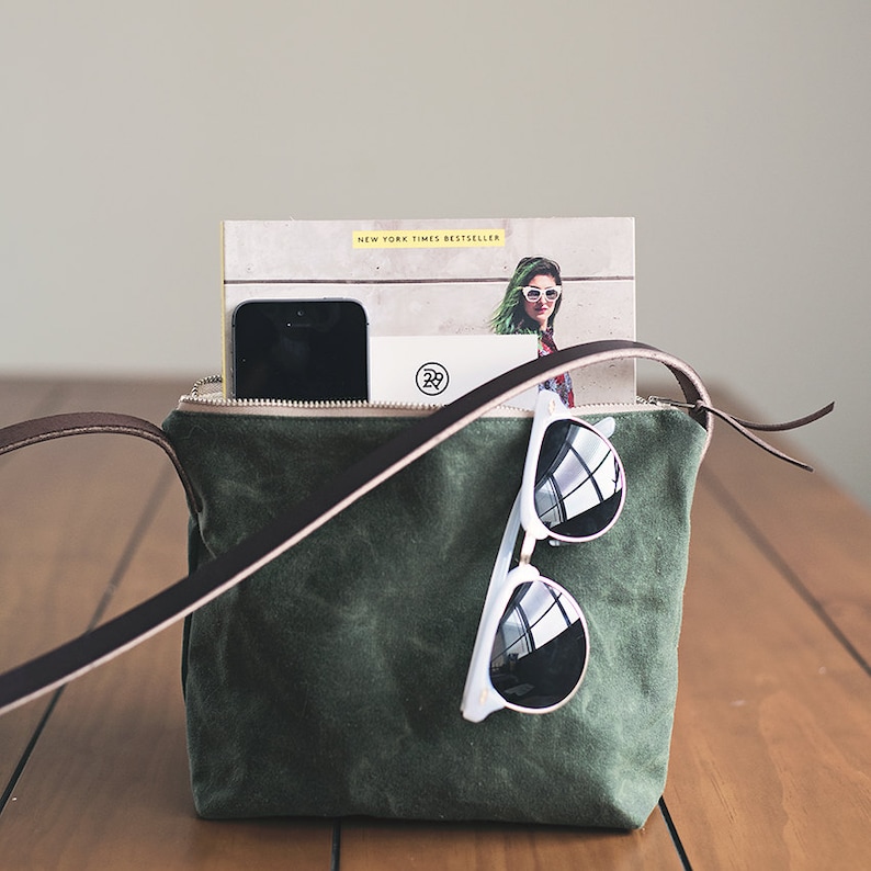 May include: A green canvas tote bag with a brown leather strap. The bag is open and contains a smartphone, sunglasses, and a book with the text "New York Times Bestseller".