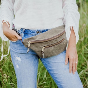 May include: A person wearing blue jeans and a white shirt with a brown canvas fanny pack with two zippered compartments and gold zipper pulls.