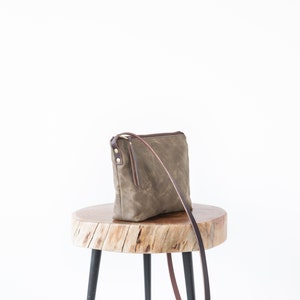 May include: A brown canvas crossbody bag with a brown leather strap and gold hardware. The bag is sitting on a wooden stool with black legs.