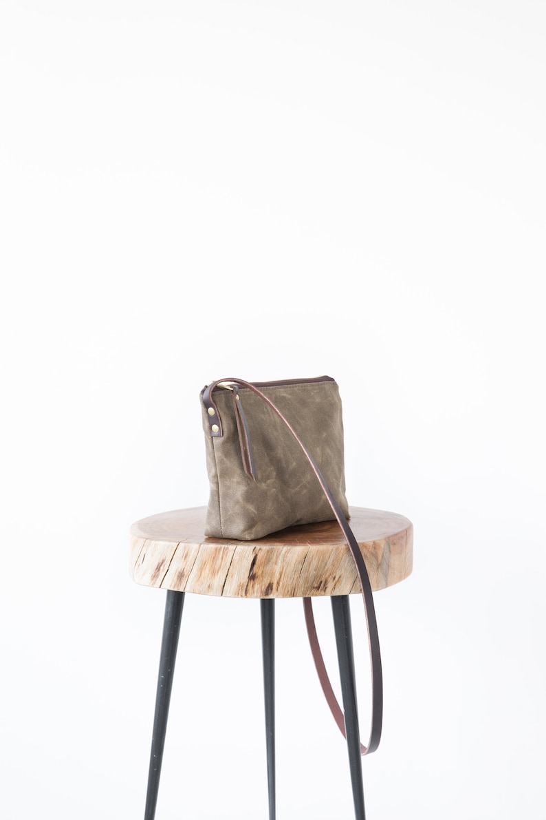 May include: A brown canvas crossbody bag with a brown leather strap and gold hardware. The bag is sitting on a wooden stool with black legs.