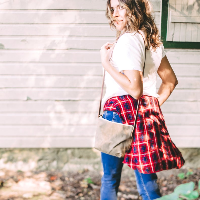 May include: A woman wearing a white t-shirt, red and black plaid shirt, and blue jeans. She is carrying a brown leather crossbody bag.