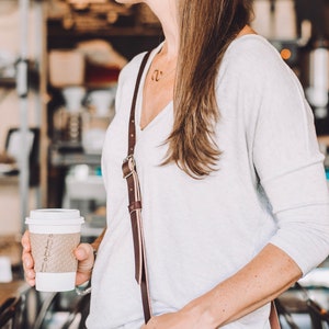 May include: A woman wearing a white long-sleeved shirt with a brown leather strap. She is holding a white to-go coffee cup with brown text that says "Brown Paper Bag".